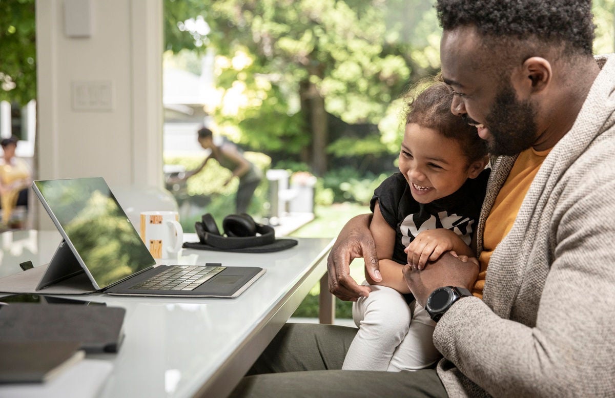 A person works at a table with a laptop while holding a young child on their lap, with a backyard  visible outside.
