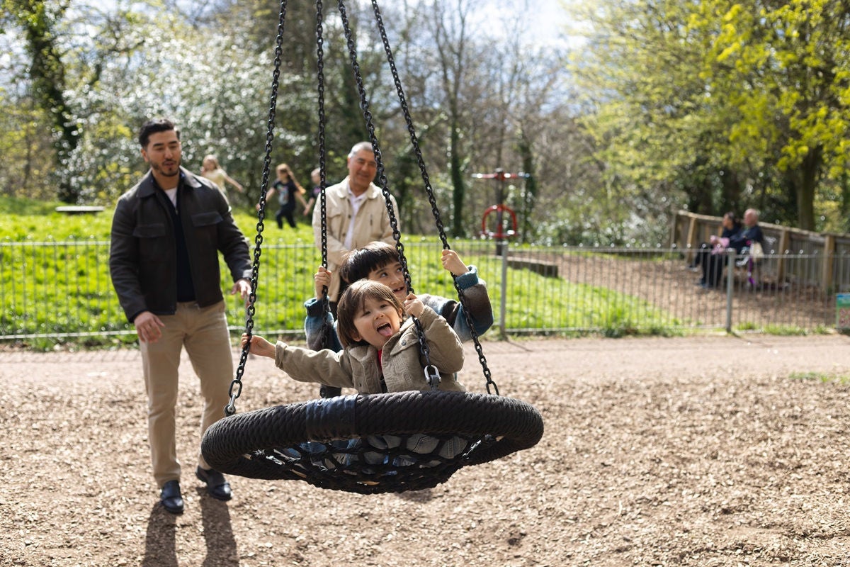Adults stand near a playground swing as a child rides a circular net swing in a sunny park with trees and a fenced play area in the background