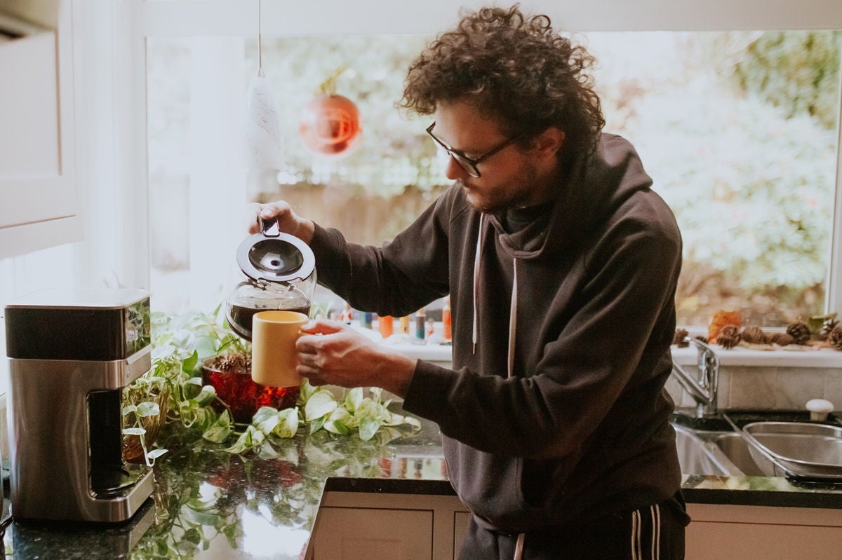 A person in a cozy kitchen pouring freshly brewed coffee into a yellow mug beside a coffee maker.