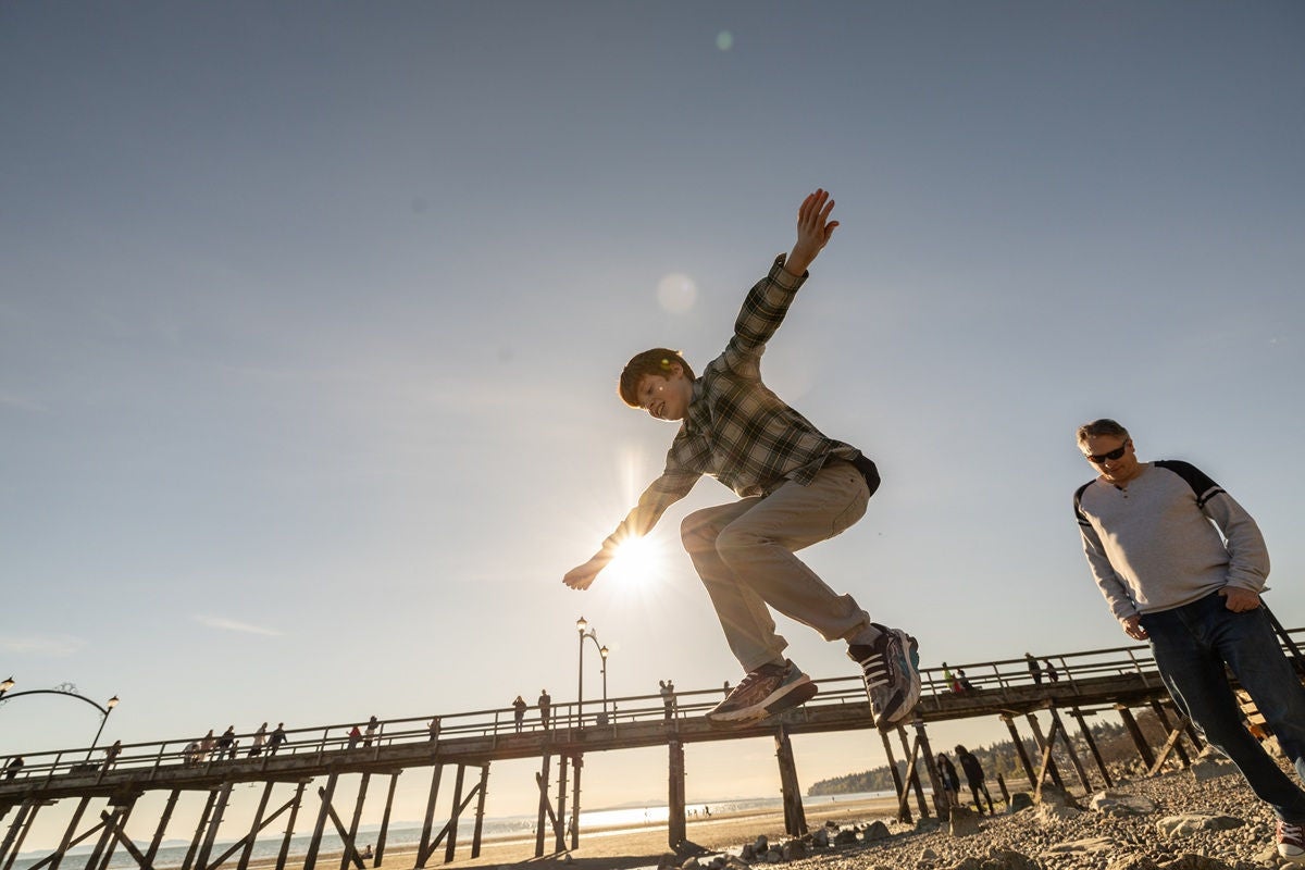 A child jumps across rocky beach stones near a wooden pier, captured from a low angle against a wide sky, while an adult walks a short distance behind along the shoreline in warm evening light.