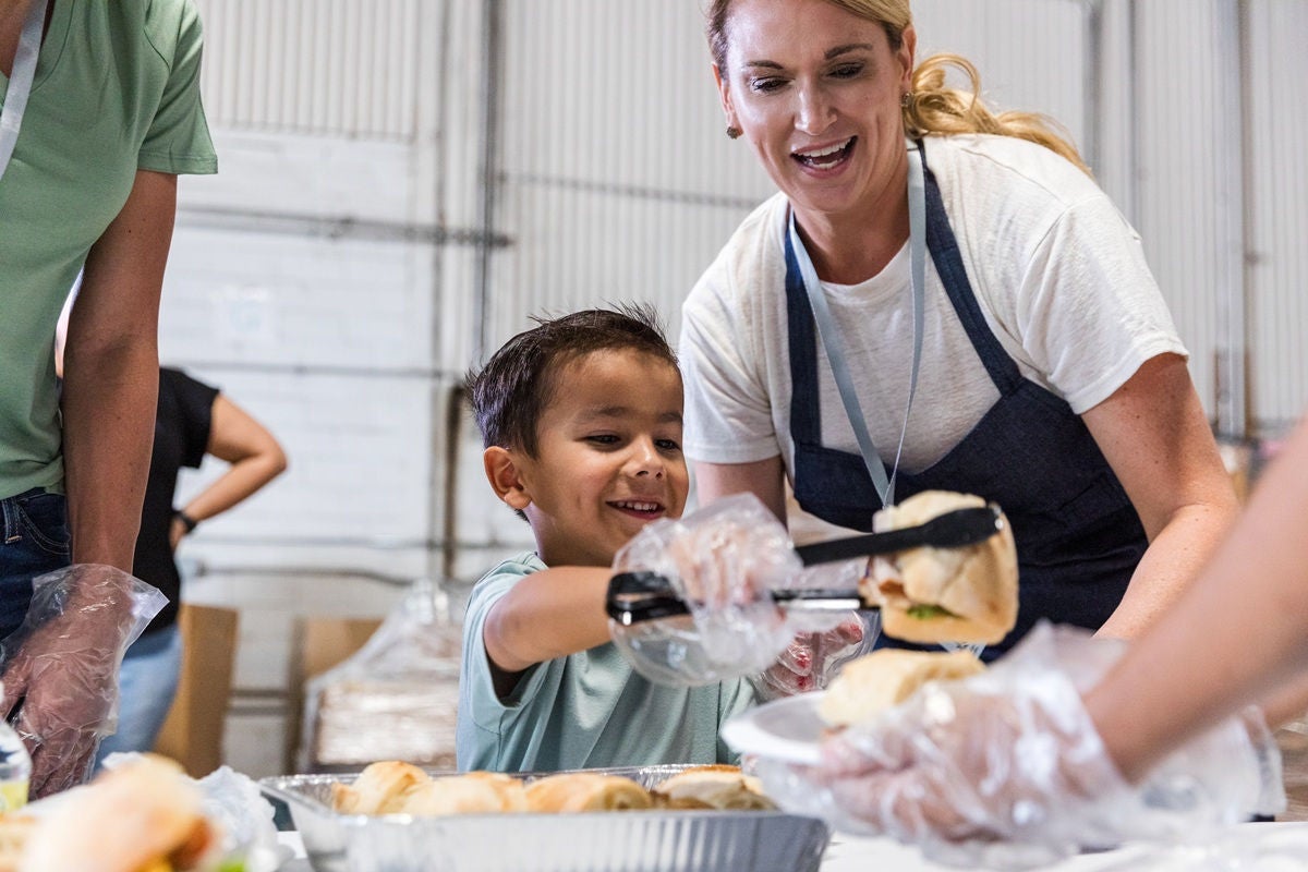 A child and an adult serve food together at a community meal, placing sandwiches into trays alongside other volunteers.