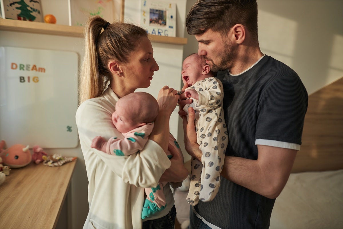 two adults stand in a nursery holding newborn twins, comforting one baby who is crying while the other rests quietly in their arms.
