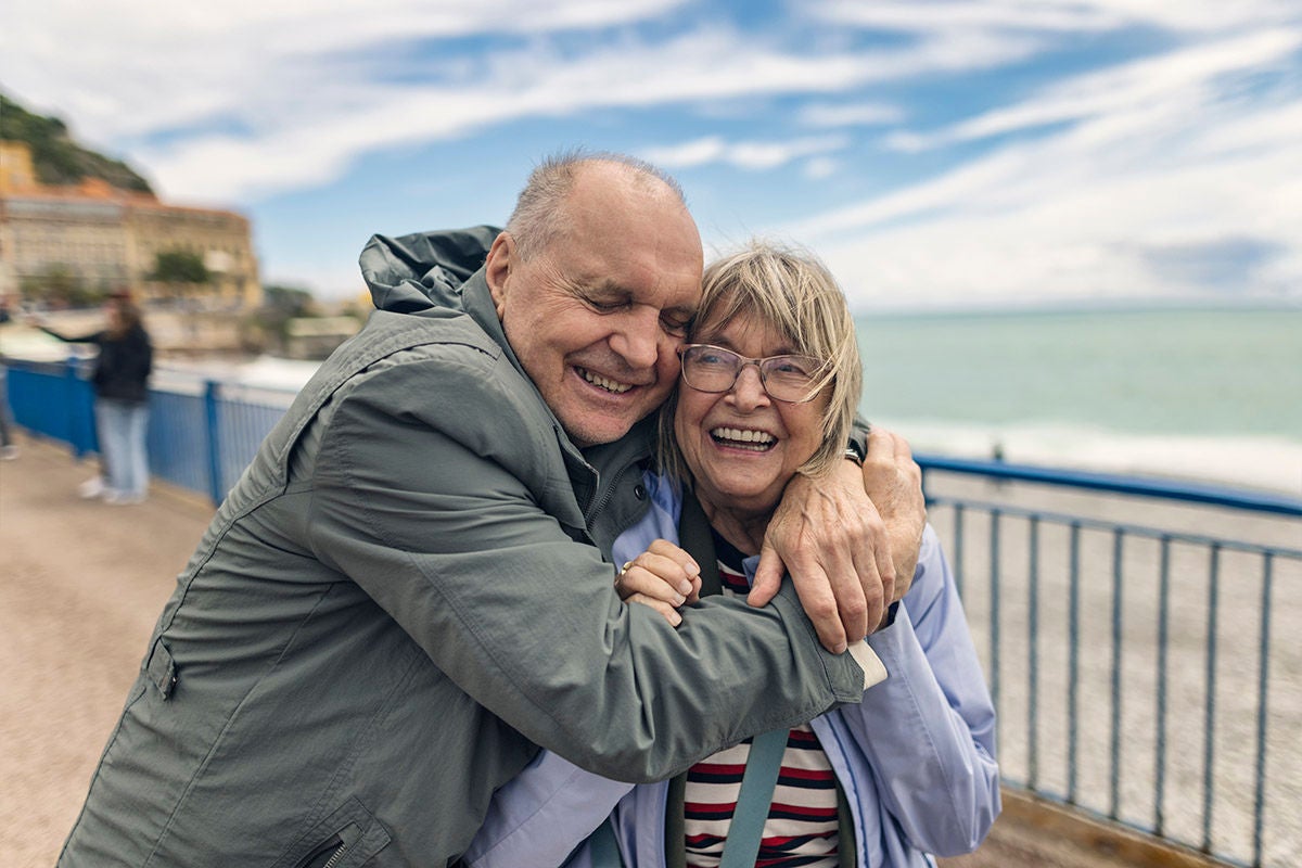 A smiling elderly couple is embracing on a seaside promenade in Nice (Promenade des Anglais) , sharing a joyful moment. The background features a blue railing and the sea under a partly cloudy sky.
Shot with Canon R5