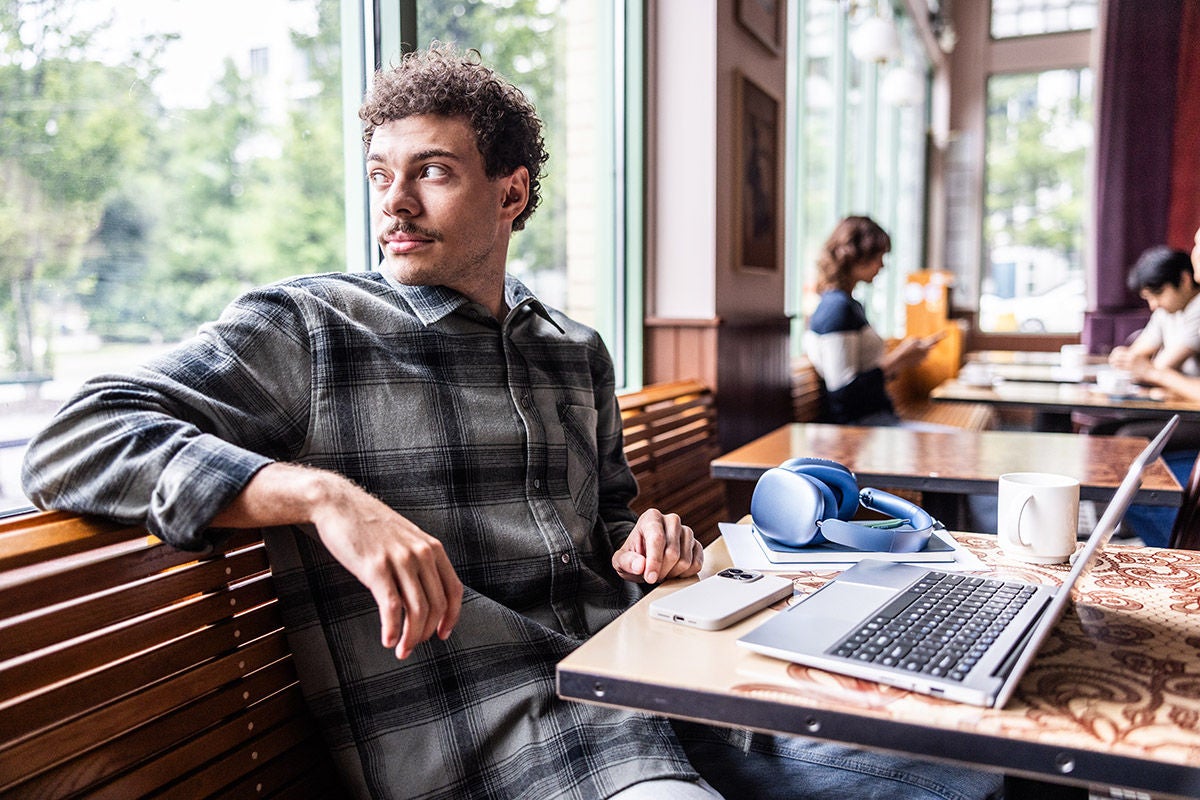 Person sitting at a café table by a window with a laptop, smartphone, and headphones, relaxing while working or browsing in a coffee shop setting.
