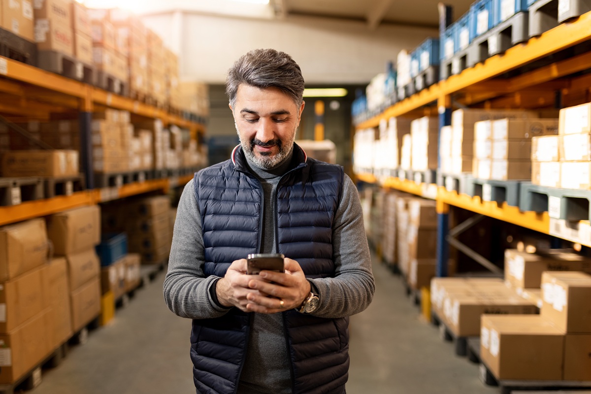 A person stands in a warehouse aisle checking a smartphone, surrounded by shelves stacked with labeled boxes and inventory.