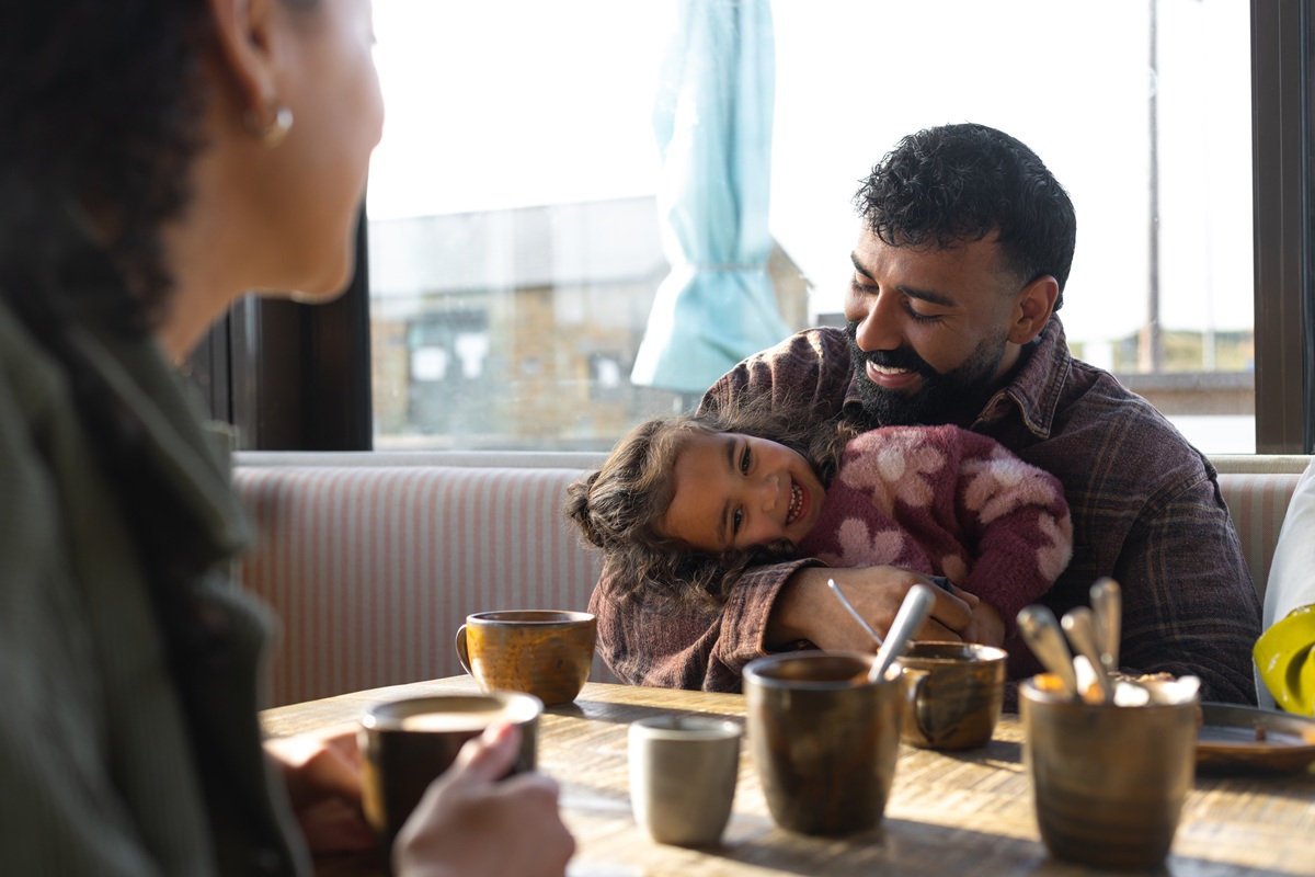 A family sitting at a café table, sharing drinks while one adult holds a child, creating a warm, intimate moment.